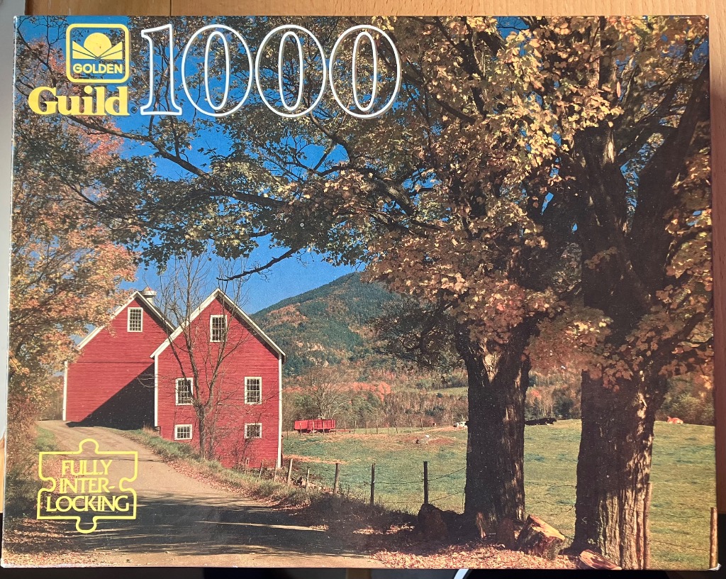 A pair of red barns along a country road with large trees and mountains in the background during autumn.