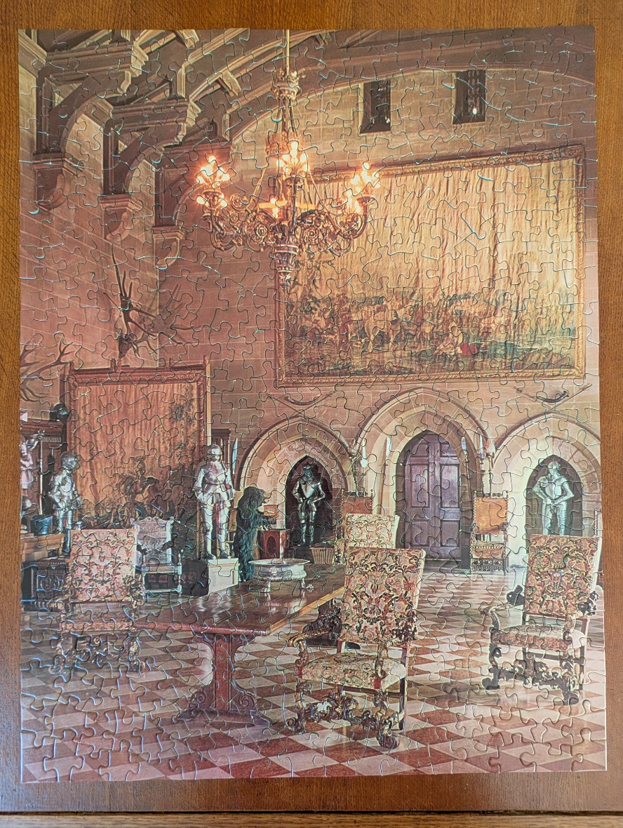 An opulent castle hall featuring suits of armor, ornate furniture, a large chandelier, and a decorative wall tapestry.