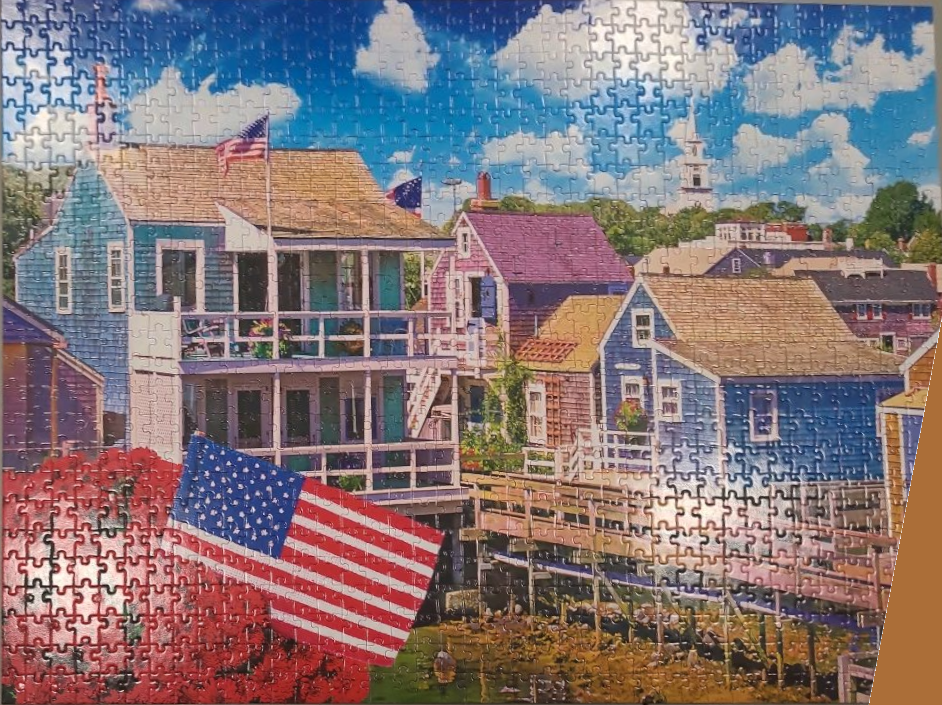 A coastal village scene featuring colorful houses, American flags, and a blue sky with clouds.