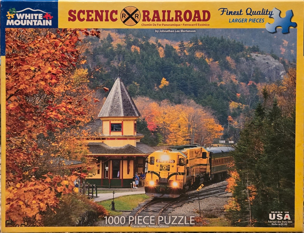 A yellow diesel locomotive passing a vintage Victorian-style train station surrounded by vibrant autumn foliage.