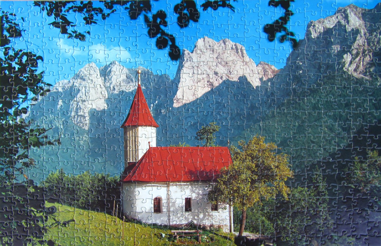 A small white church with a bright red roof nestled in front of towering, craggy mountains.