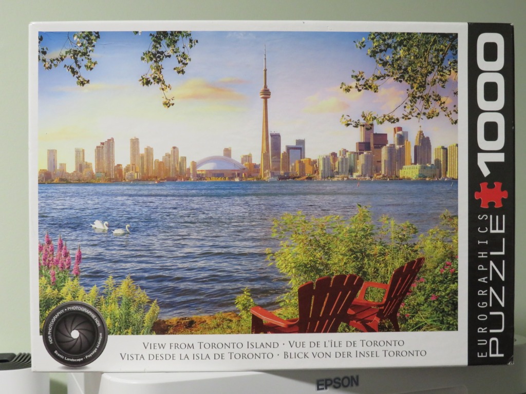 A scenic photograph of the Toronto skyline across the water as viewed from a park with two red chairs and white swans.