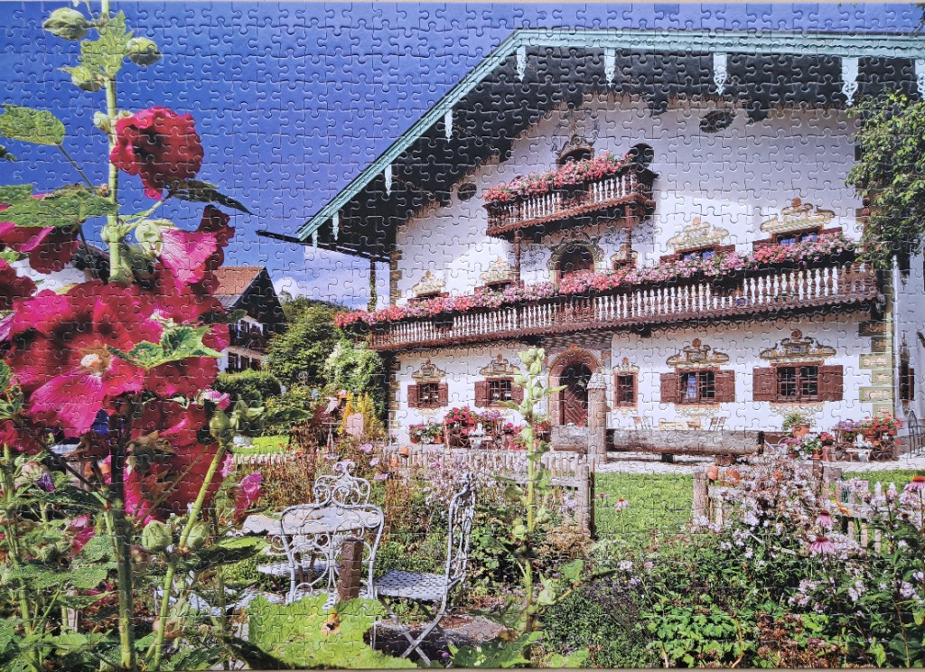 A sunny, traditional white Alpine farmhouse with flower-filled balconies, framed by tall vibrant red hollyhocks.