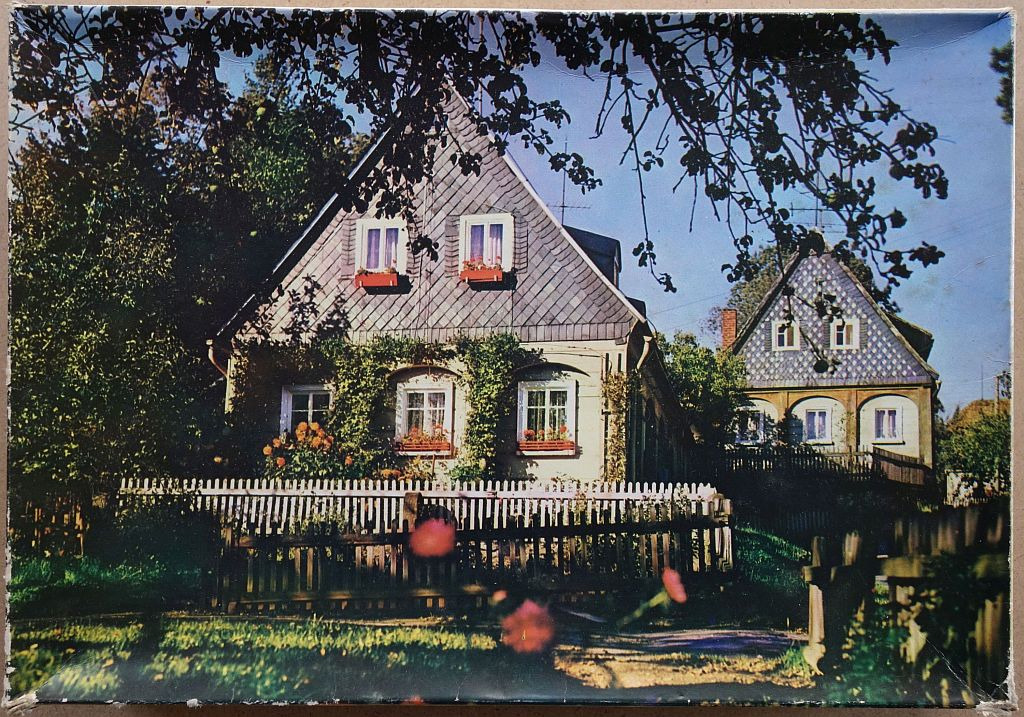 A vintage-style photograph of two slate-tiled houses behind a wooden fence with leafy trees overhead.