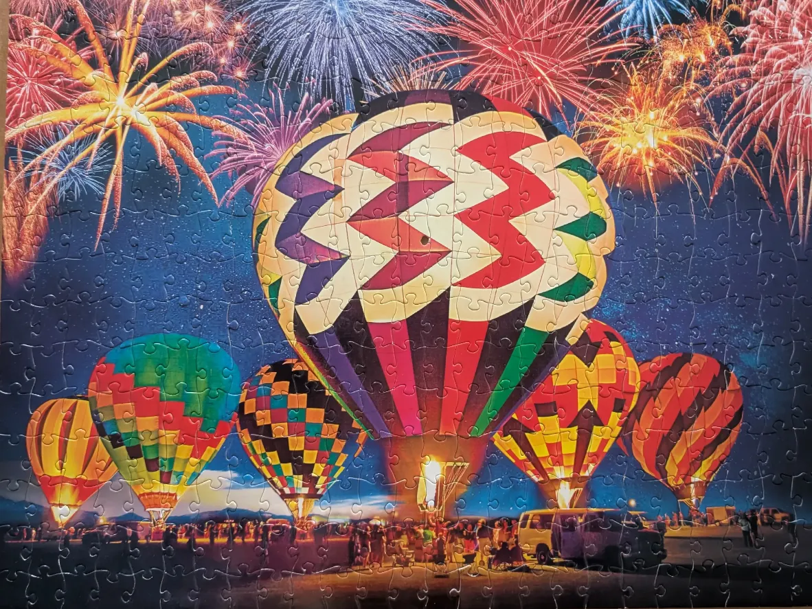 Multiple hot air balloons lit from within against a night sky filled with fireworks.