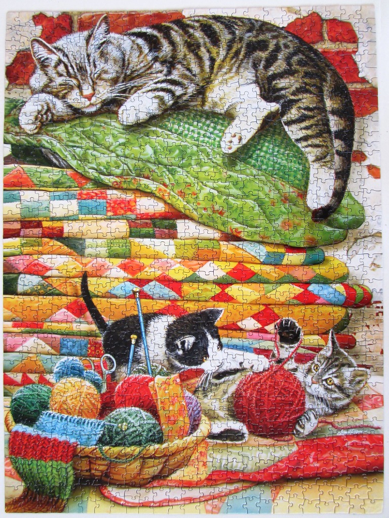 A tabby cat sleeping on quilts above two kittens playing with yarn in a basket.