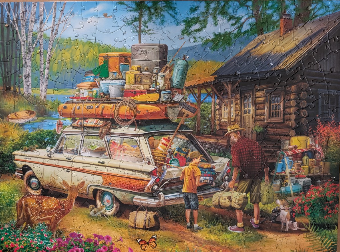 A family loading a vintage station wagon packed with camping gear in front of a rustic log cabin by a lake.