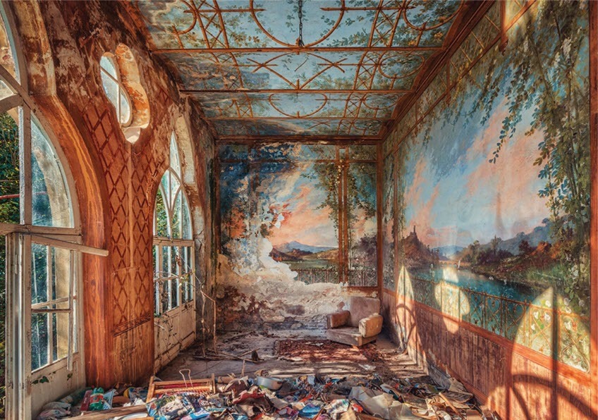 An abandoned room with peeling floral wallpaper murals, a single armchair, and sunlight streaming through arched windows.