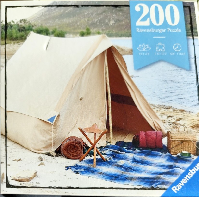 A tan canvas tent set up on a sandy shore by a lake, accompanied by a small tripod stool, blankets, and a wicker basket.
