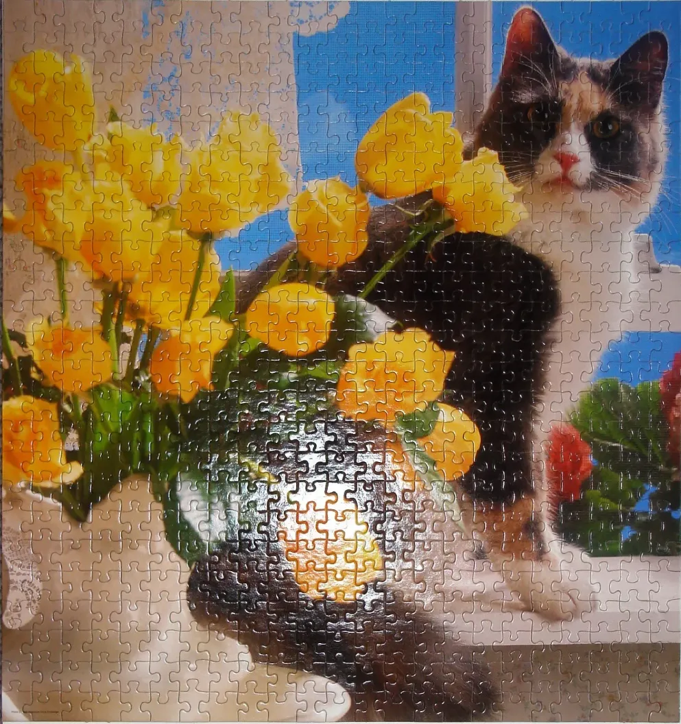 A calico cat sitting on a windowsill next to a vase of yellow tulips against a bright blue sky.