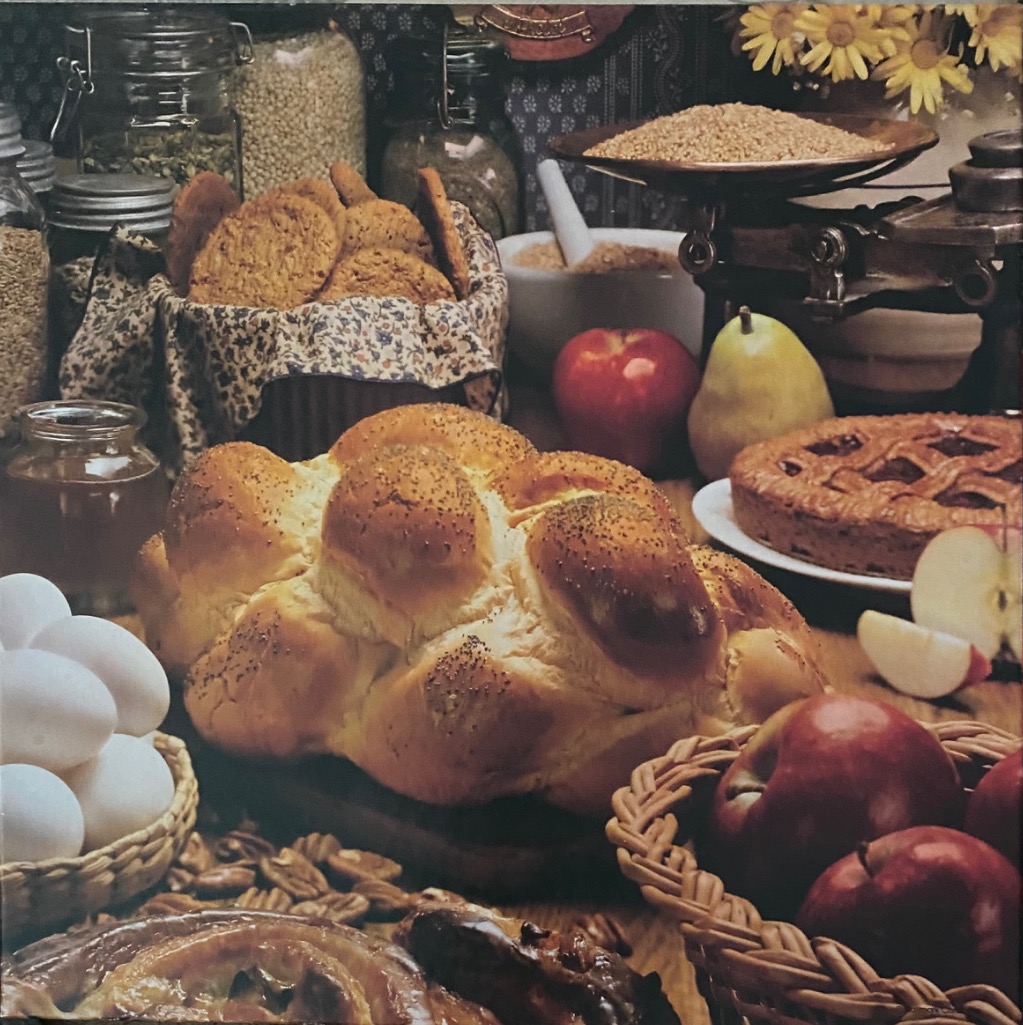 A still life arrangement featuring a golden braided loaf of bread, various fruits, and rustic kitchen ingredients.