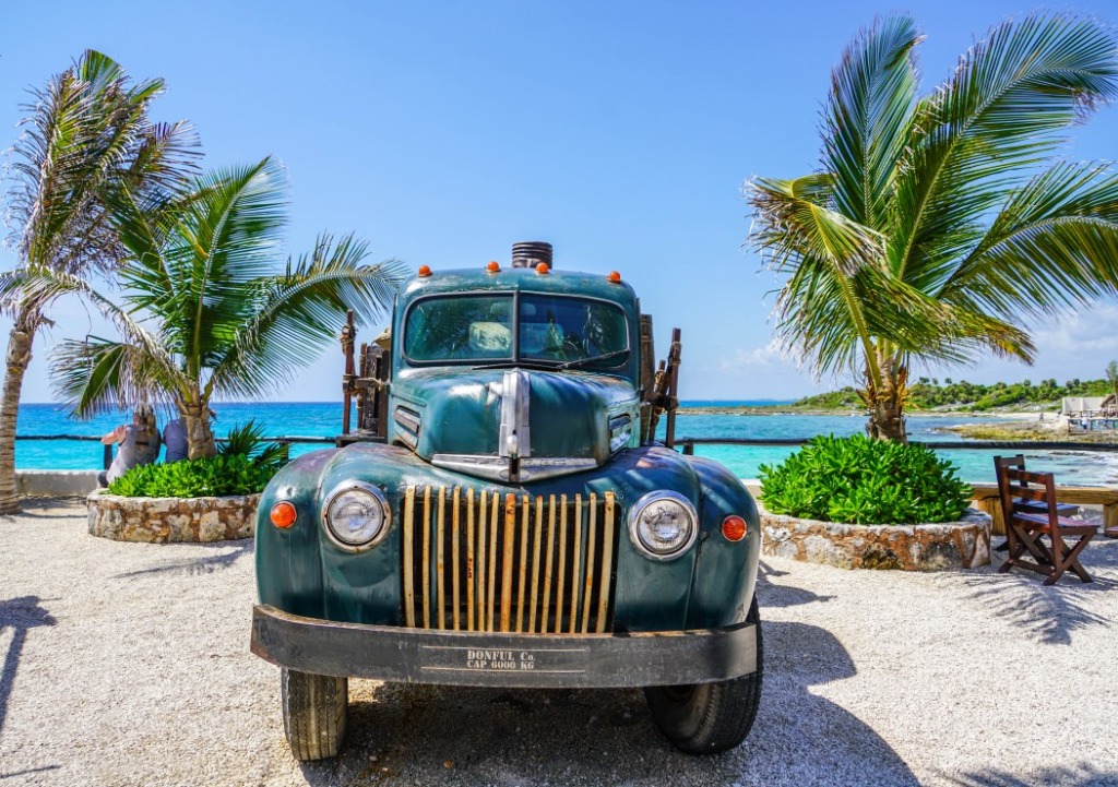 An old, dark green vintage truck parked on a gravel path overlooking a turquoise Caribbean sea with palm trees.