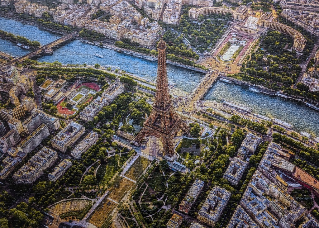 An aerial view of the Eiffel Tower and the surrounding Parisian landscape, including the Seine River and city blocks.