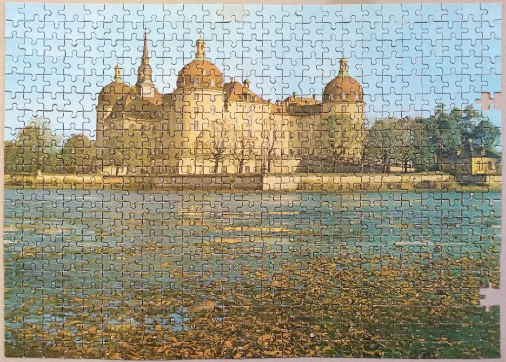 A photograph of Moritzburg Castle reflected in a pond covered with lily pads under a clear blue sky.