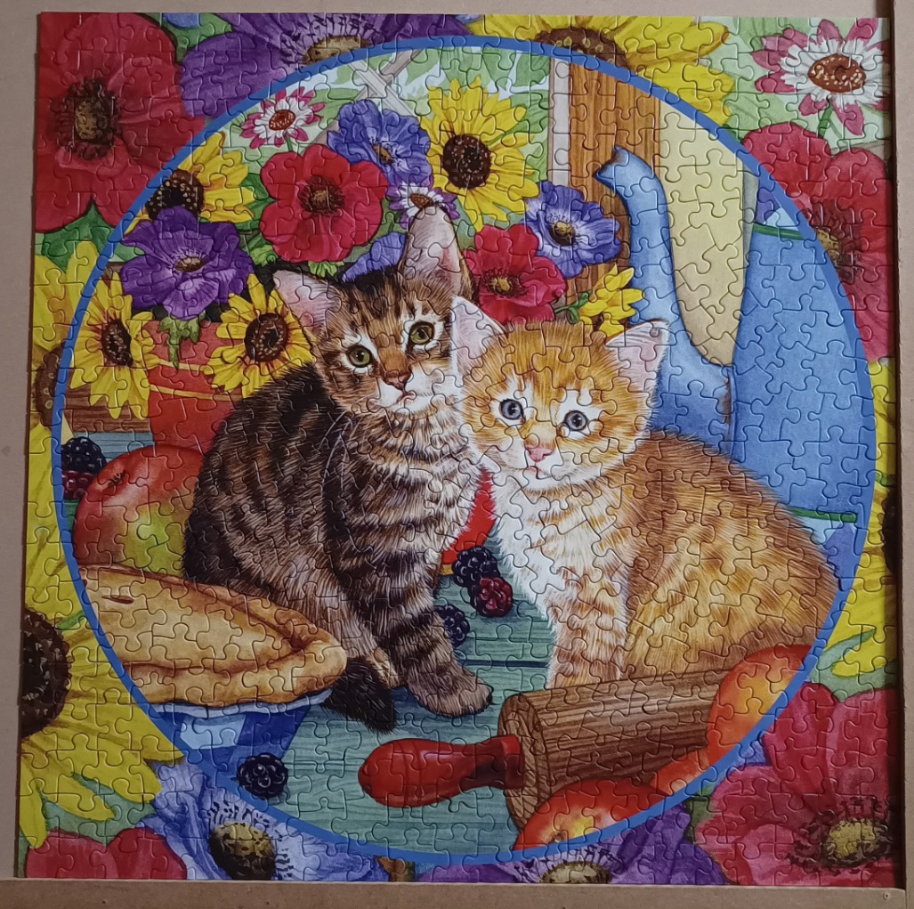 Two kittens sitting on a blue wooden surface surrounded by vibrant flowers, a pie, and a rolling pin.