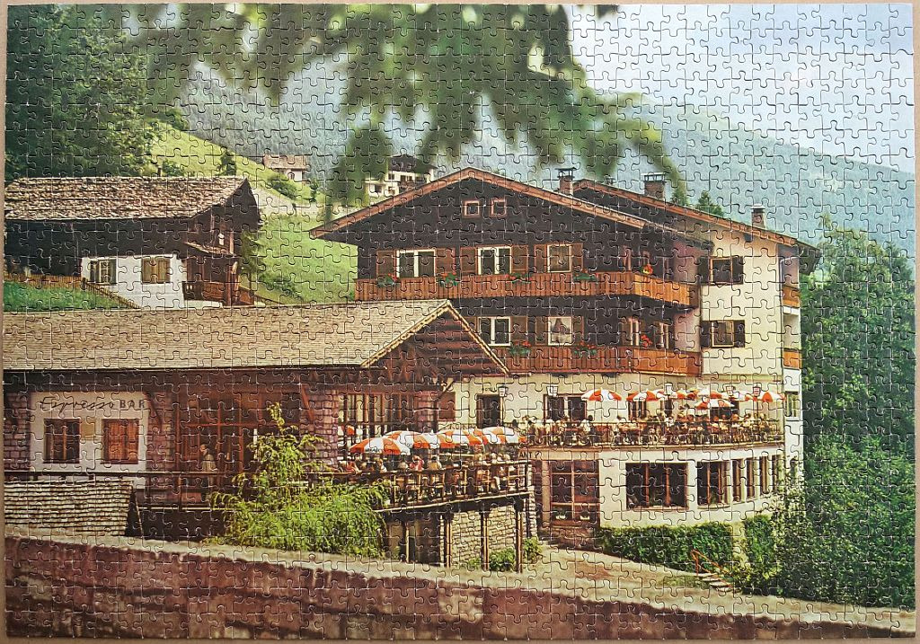 A European alpine hotel with an outdoor terrace and espresso bar, surrounded by lush green hills and trees.