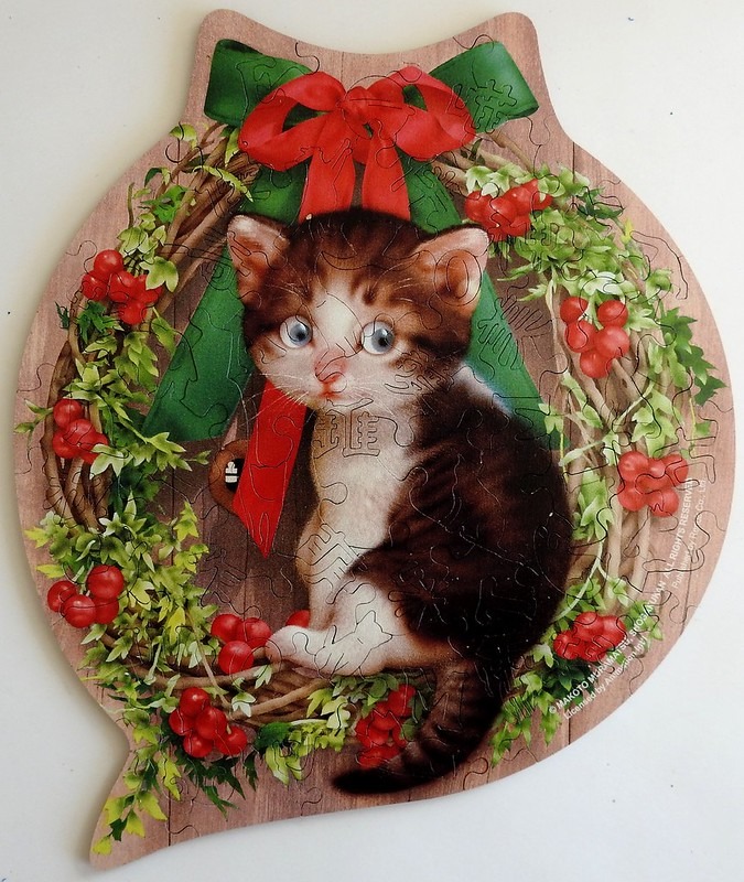 A cute brown tabby kitten sitting inside a Christmas wreath against a wood-grain background.