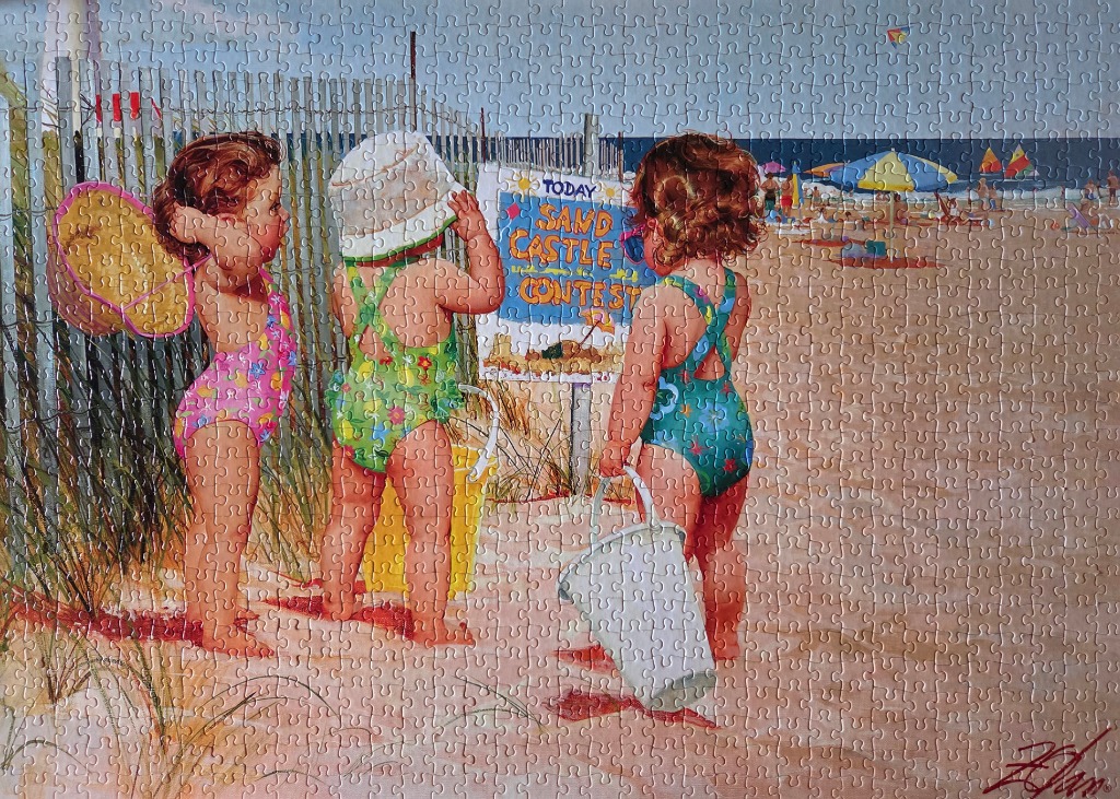 Three toddlers standing on a beach in front of a sandcastle contest sign.