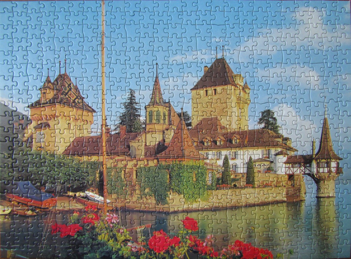 A picturesque European castle with stone walls and red roofs sits on the shore of a lake, framed by red geraniums.