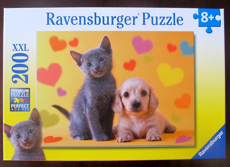 A grey kitten and a light brown puppy sitting side-by-side against a yellow background with colorful hearts.