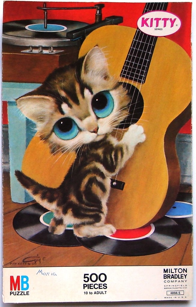 A wide-eyed tabby kitten standing next to a large acoustic guitar near a vintage record player.