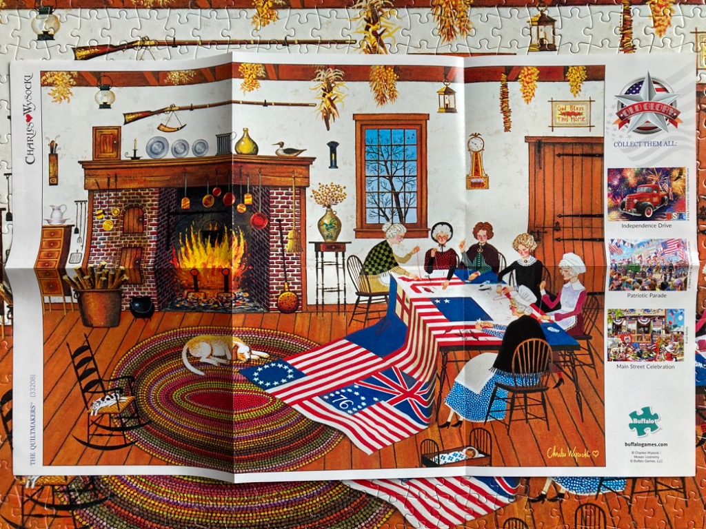 A group of women in colonial-style clothing gathered around a table sewing a large American flag.