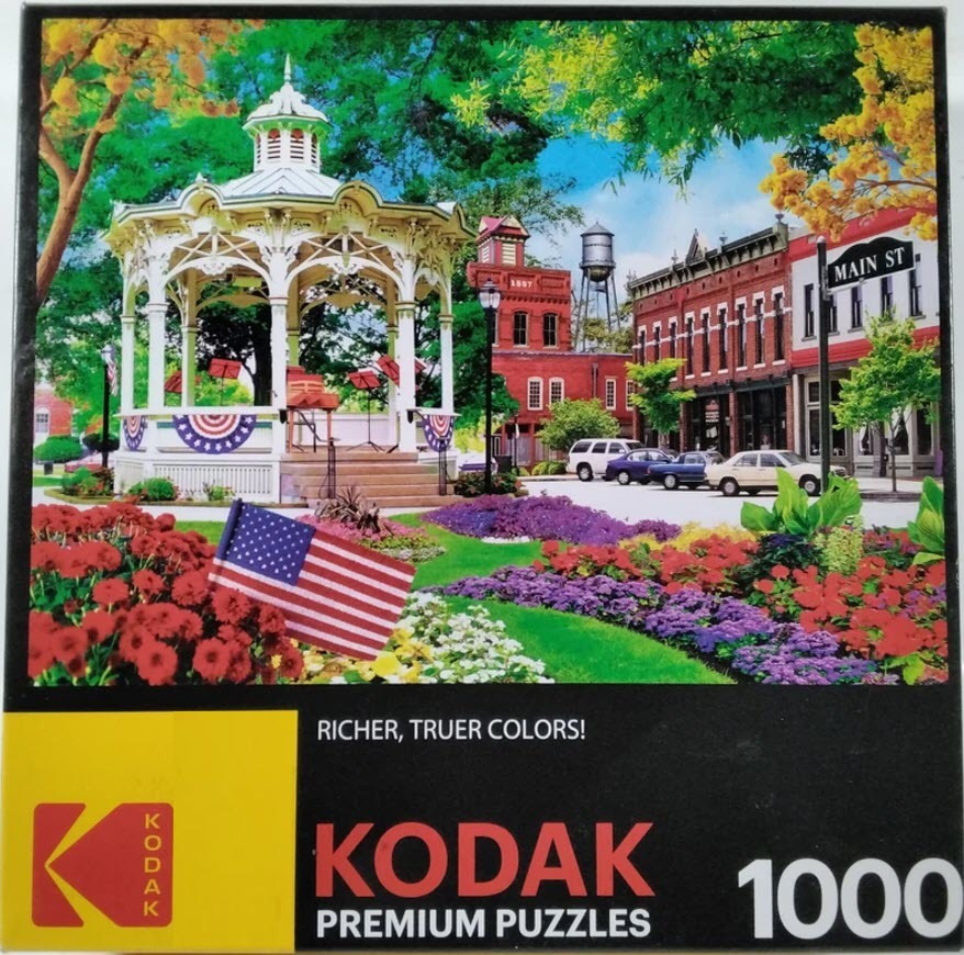 A small-town scene with a gazebo, brick buildings, and flower beds under a sunny sky.