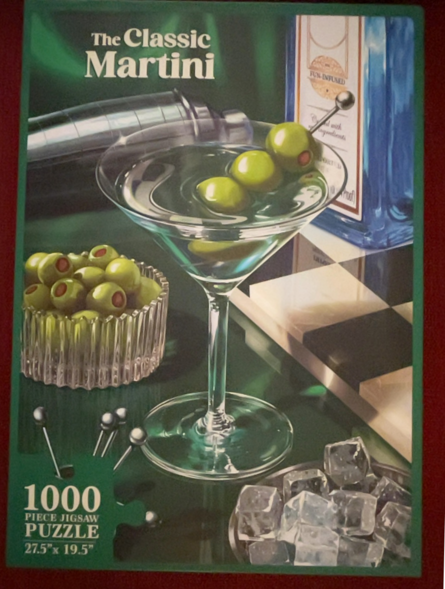 A martini glass with olives, a shaker, and a gin bottle on a checkerboard surface.