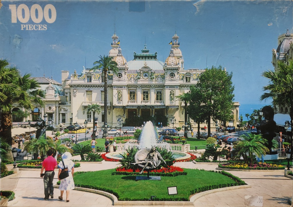 A sunny, eye-level photograph of the Monte Carlo Casino in Monaco with a fountain and park in the foreground.