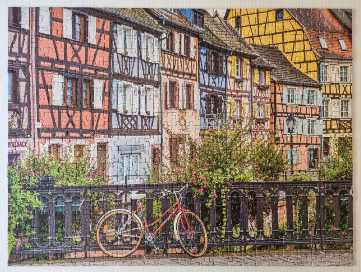 A red bicycle leaning against a black iron railing in front of colorful half-timbered buildings in Alsace, France.