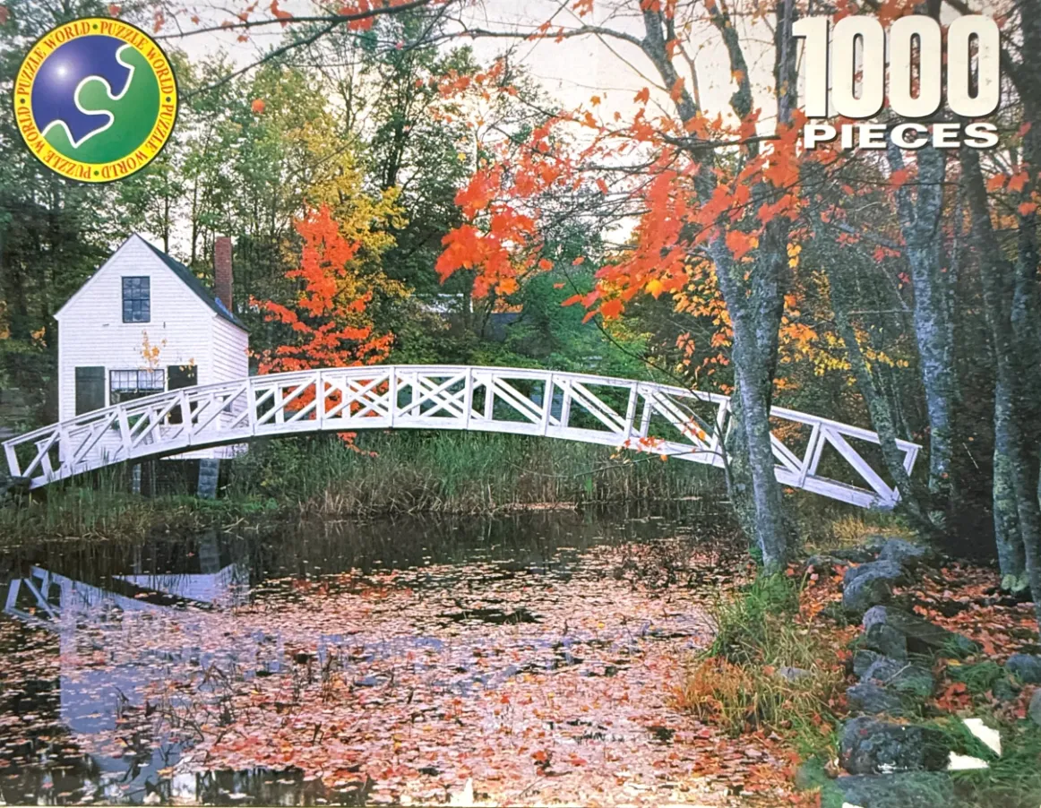 A white arched bridge crosses a stream covered in autumn leaves near a white house surrounded by fall foliage.