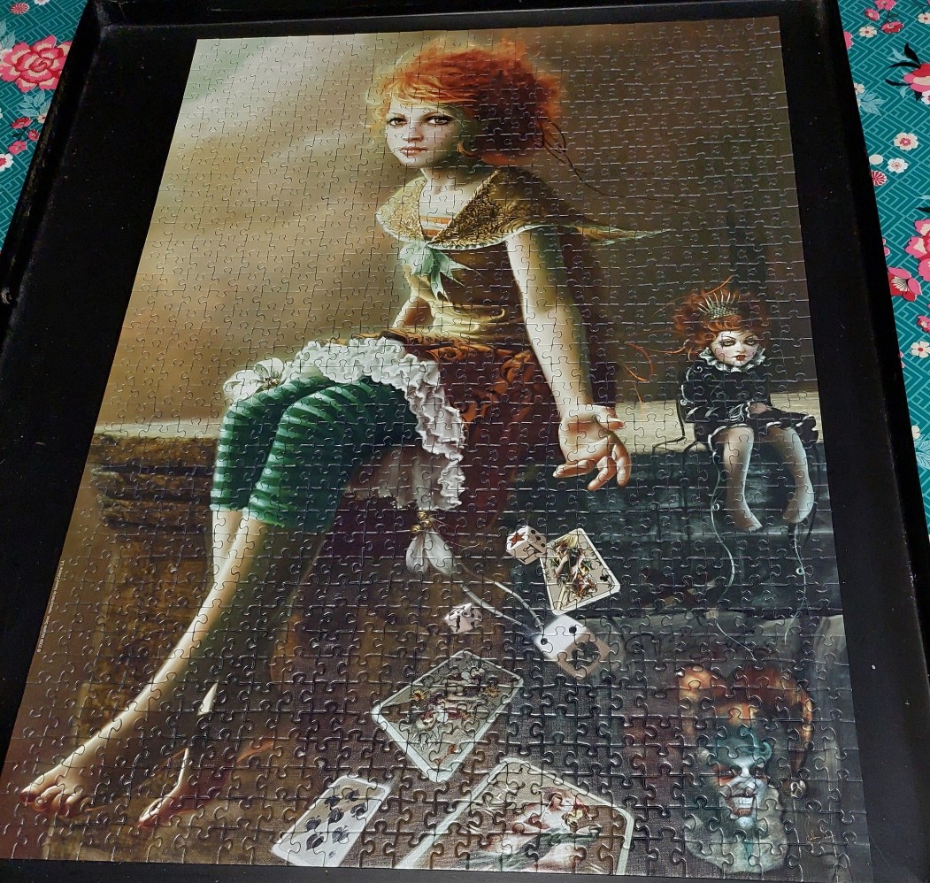 An ethereal, auburn-haired girl sits on a stone ledge next to a doll, surrounded by scattered playing cards and dice.