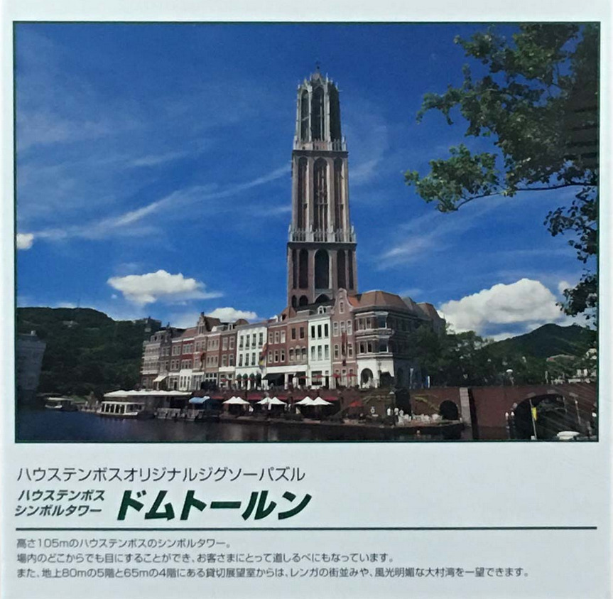 The Domtoren tower at Huis Ten Bosch rising above Dutch-style buildings and a canal under a bright blue sky.
