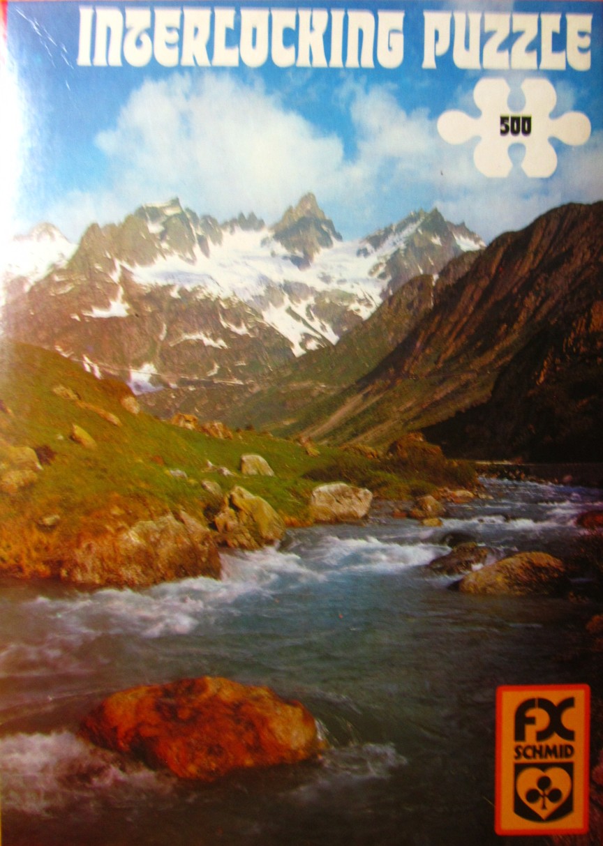 A photograph of a mountain range featuring snowy peaks above a grassy valley with a rushing river in the foreground.