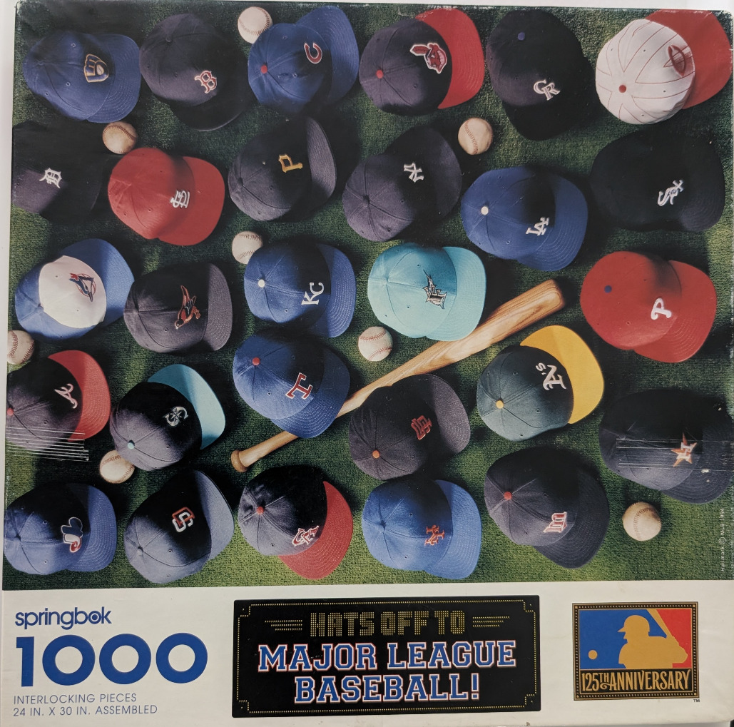 An overhead view of numerous Major League Baseball team caps arranged on green turf with baseballs and a wooden bat.