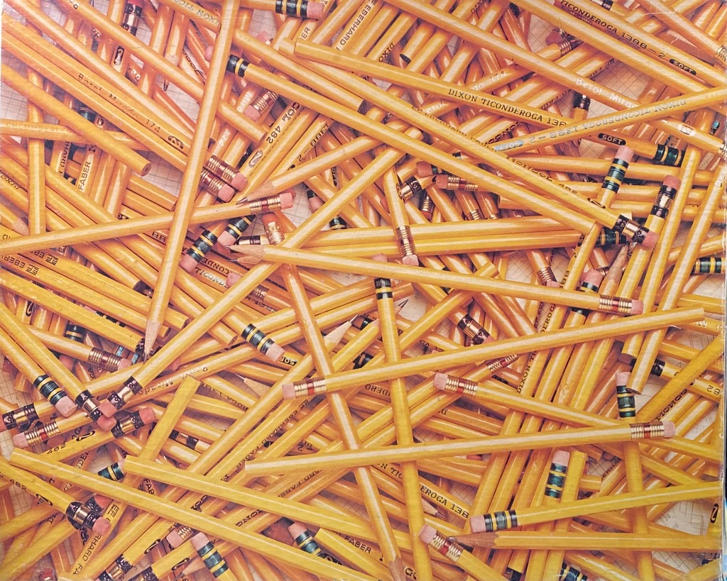 A chaotic, high-angle composition of numerous yellow pencils with pink erasers scattered over a grid-patterned surface.