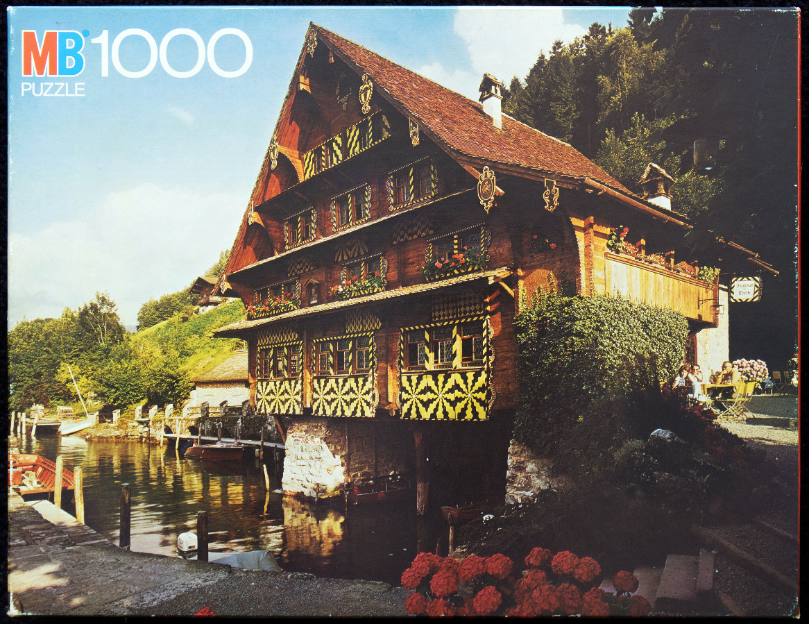 A traditional Swiss-style wooden house on the edge of a calm lake with lush green hills in the background.