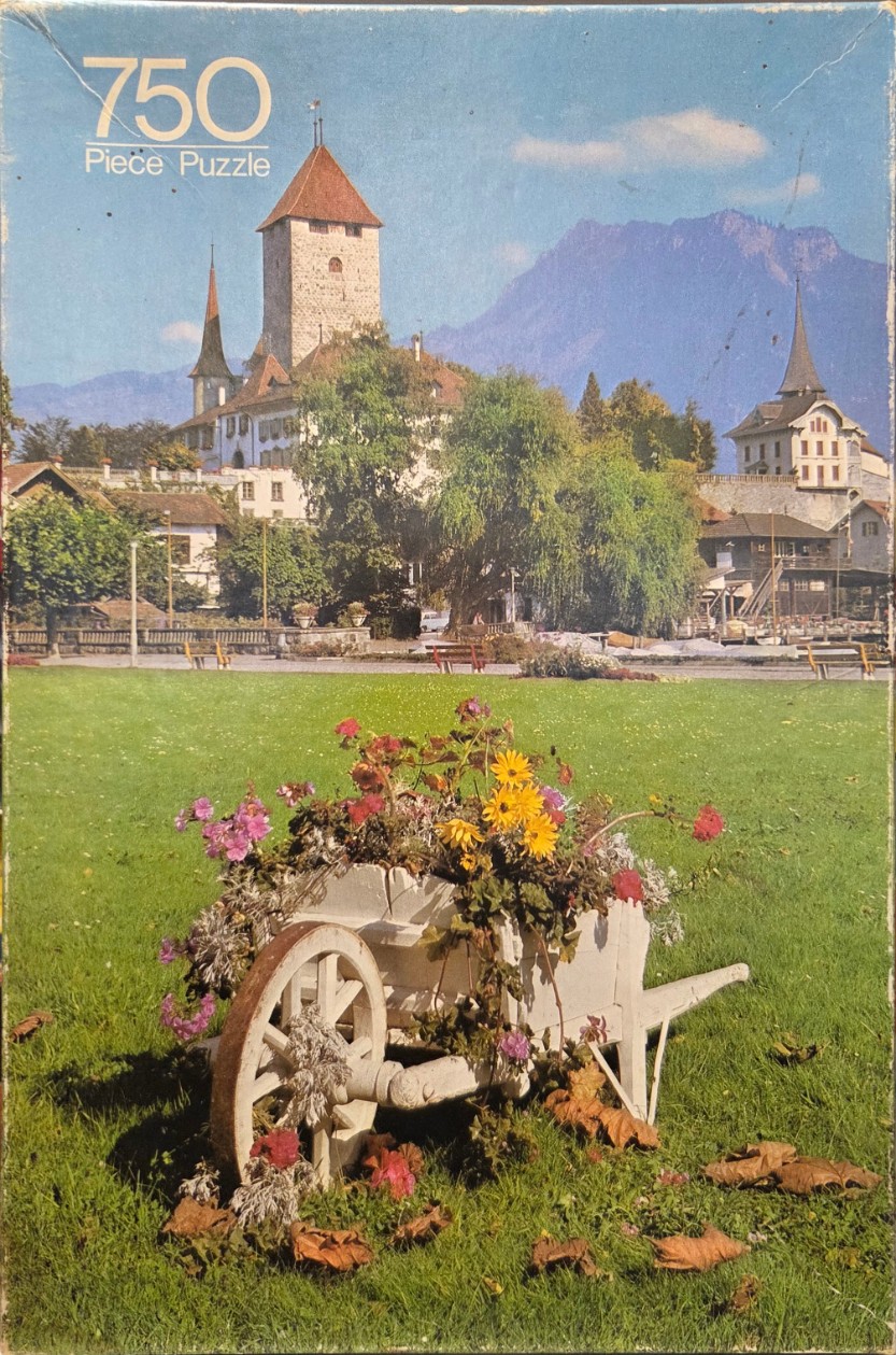 A sunny park scene with a flower-filled white wooden wheelbarrow in the foreground and a Swiss castle in the background.