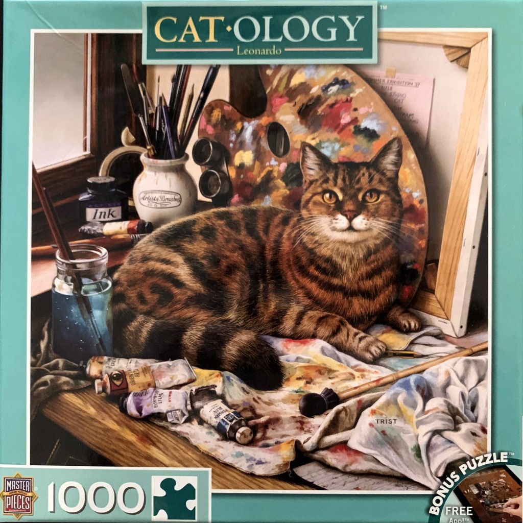 A brown tabby cat lounging on an artist's workspace covered in paint tubes, brushes, and a palette.