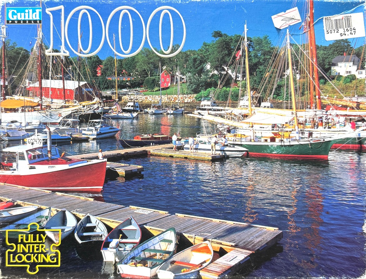 A sunny harbor scene featuring sailboats and smaller rowing boats docked along wooden piers, with red buildings in the back.