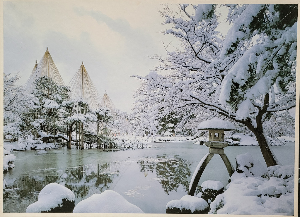 A peaceful, snow-covered Japanese garden in winter with yukitsuri ropes on trees, a stone lantern, and a calm pond.