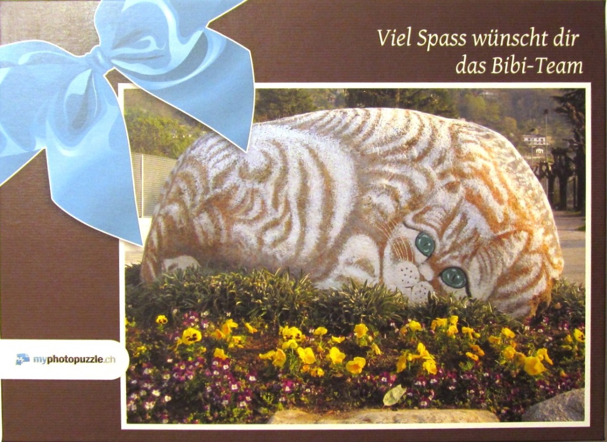 A topiary or rock sculpture shaped like a ginger tabby cat resting in a flower bed, decorated with a blue gift bow.