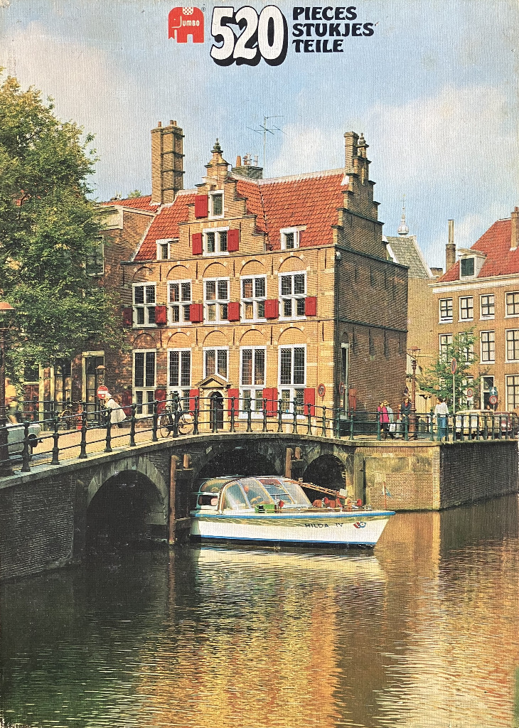 A scenic view of a brick Dutch house with red shutters situated along a canal with a small boat parked underneath a bridge.