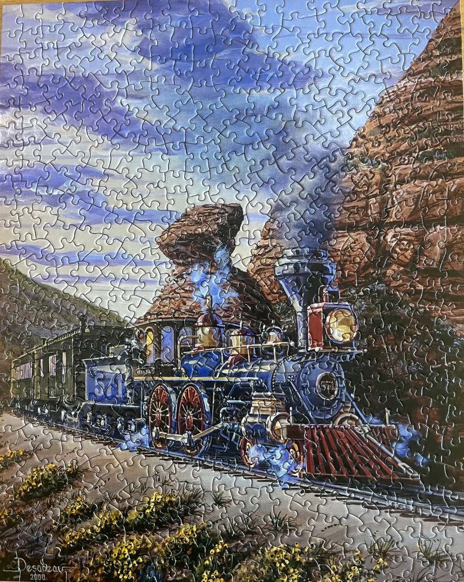 A vintage steam locomotive traveling through a rugged red rock canyon under a blue cloudy sky.