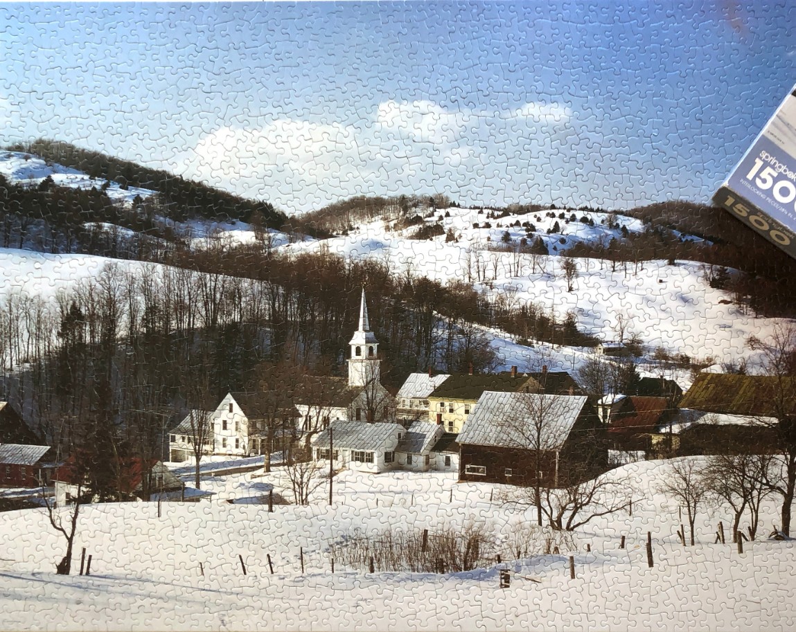A snowy New England village nestled in rolling hills under a clear blue sky.