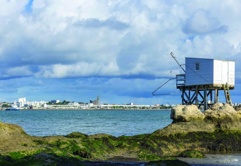 A traditional stilt fishing cabin overlooks a calm bay with a coastal town in the distance under a cloudy sky.