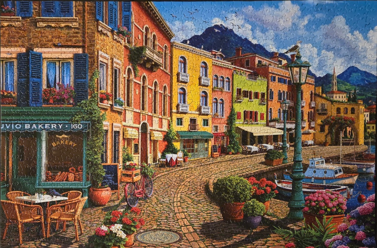 A charming Mediterranean waterfront village scene with colourful buildings, a cobblestone street, and boats.
