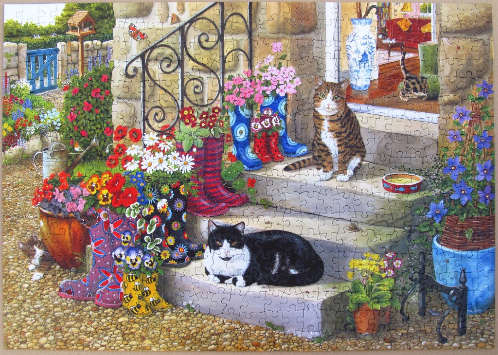 A cozy country garden scene featuring two cats, colorful rain boots used as planters, and a stone patio.