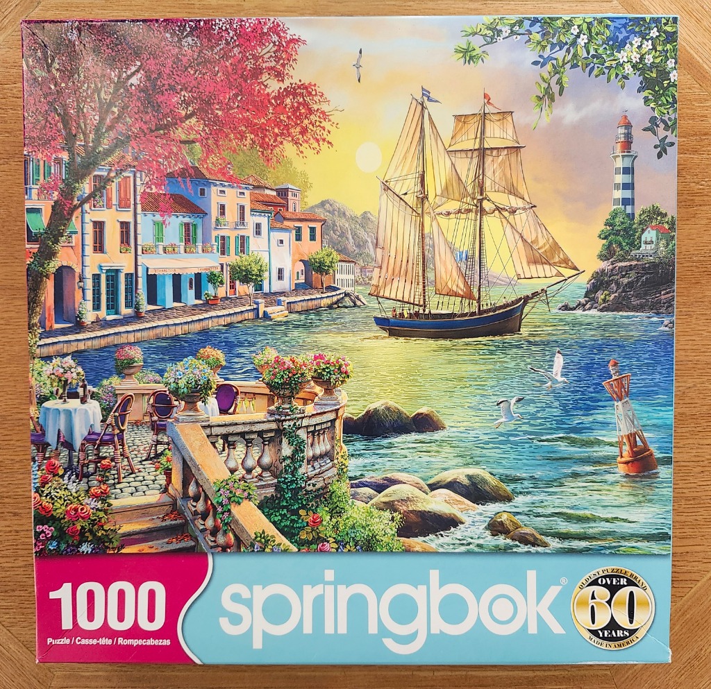 A peaceful coastal town scene with a sailboat, a lighthouse, and a flower-filled terrace overlooking the water at sunset.