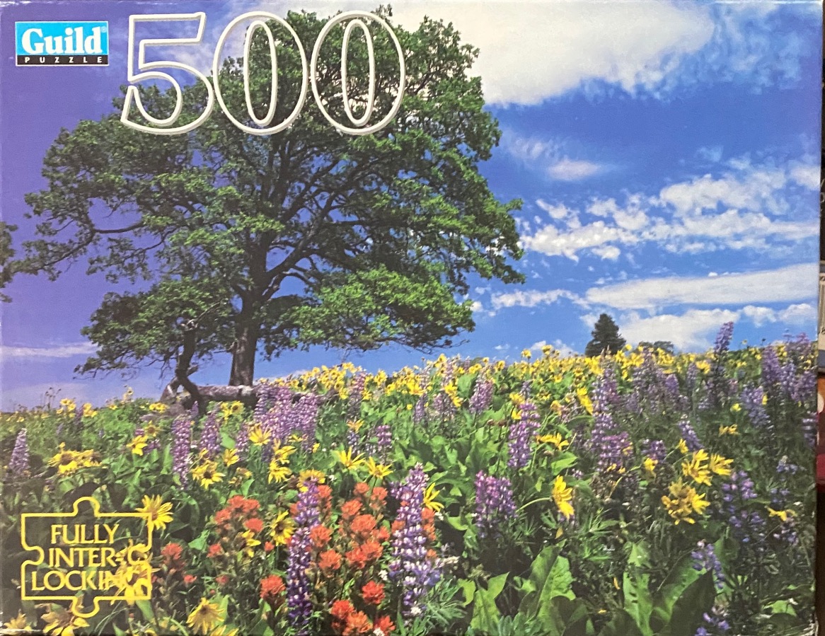 A large, leafy tree stands in a field of colorful wildflowers under a bright blue sky.